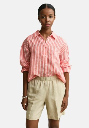 Vrouw draagt een koraalroze en wit gestreept overhemd met opgerolde mouwen en beige shorts met hoge taille, staand tegen een effen achtergrond.