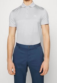 Man i ljusgrå kortärmad polotröja med subtilt strukturerad mönster och blå byxor mot en enfärgad bakgrund.