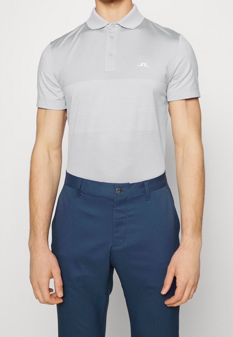 Man i ljusgrå kortärmad polotröja med subtilt strukturerad mönster och blå byxor mot en enfärgad bakgrund.