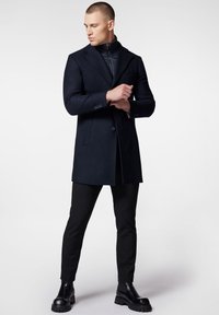 Granatowy płaszcz z wełny w kształcie strukturalnym, z klapami w kształcie notcha, zapięciem na pojedynczy guzik i dużymi kieszeniami, noszony na zapinaną na zamek wewnętrzną warstwę.