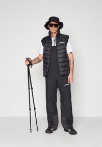 Black puffer vest over a white shirt, black overalls, black bucket hat, and sunglasses; holding trekking poles; sporty design.