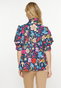Hemd mit Blumenmuster in Marineblau und Rüschensleeves. Präsentiert pinke, blaue und gelbe Blumen sowie Blätter. Leichter Stoff mit Textur.
