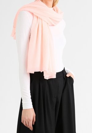 Femme portant une écharpe rose clair drapée sur un haut blanc à manches longues et un pantalon noir ample taille haute, debout avec une main dans la poche.