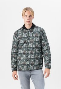Veste matelassée avec un imprimé vitrail en vert, bleu et jaune, avec un col noir et des poignets élastiques, ainsi qu'une poche avant.