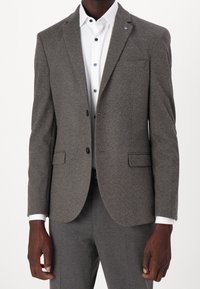 Grijze wollen blazer met een gestructureerde afwerking, sluiting met één knoop, notch lapels en voorzakken. Gecombineerd met een wit shirt met blauwe knopen.