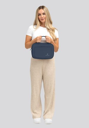 Sac en tissu bleu marine avec fermeture éclair et anse supérieure, présentant un design plat et un détail de logo à l'avant. L'utilisateur porte un pantalon texturé beige clair.