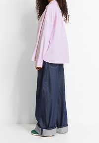 Camicia oversized a righe rosa chiaro con maniche lunghe, abbinata a jeans in denim scuro a gamba larga e calzature con dettagli verdi.