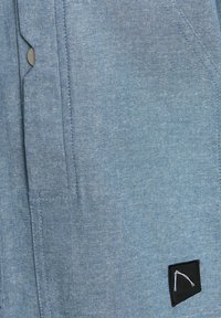 Tessuto blu con una sottile texture, caratterizzato da un'etichetta nera con un design geometrico. Include bottoni a pressione in metallo e tasche laterali.