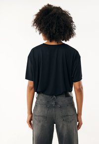 Schwarzes T-Shirt mit kurzen Ärmeln und lockerer Passform, kombiniert mit hochgeschnittenen grauen Jeans aus Denim mit Taschen auf der Rückseite und einer strukturierten Oberfläche.