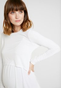 Blusa de maternidade branca de mangas longas com uma parte inferior suavemente franzida, textura lisa e decote redondo.