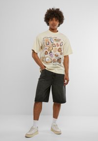 Off-white grafisk t-skjorte med fargerik agatmønster, kombinert med mørk denimshorts. Modellen har på seg hvite sneaks og synlige ankelstrømper.