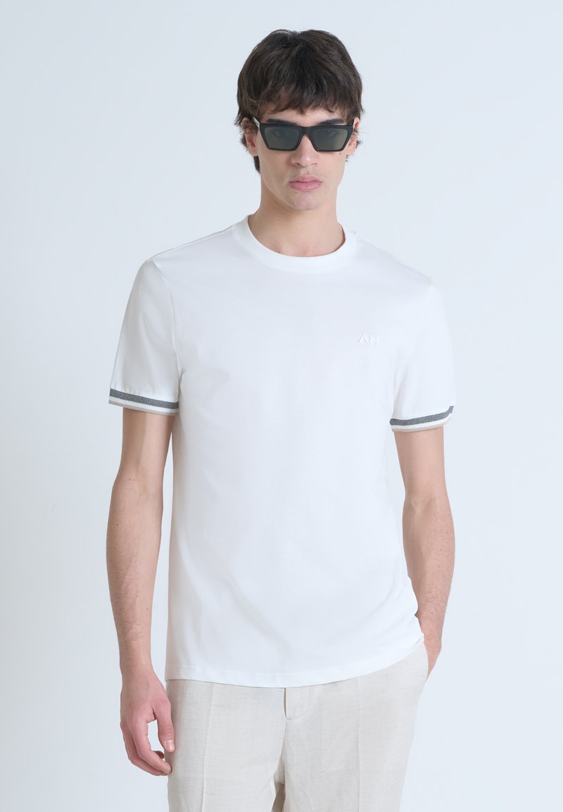 Camiseta blanca de manga corta con acentos de rayas grises en las mangas, hecha de una tela suave, combinada con gafas de sol oscuras y pantalones claros.