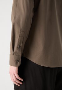 Camisa castanha de mangas longas com uma textura suave, fecho com botão nos punhos e uma bainha curva. A camisa é combinada com um tecido escuro embaixo.