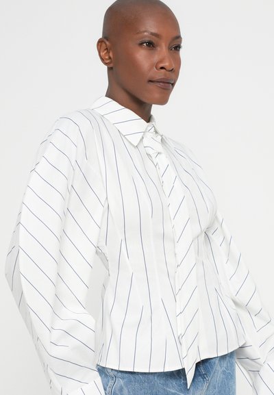 Person with shaved head wearing a white blouse with blue vertical stripes and a tied collar, paired with light blue denim jeans.