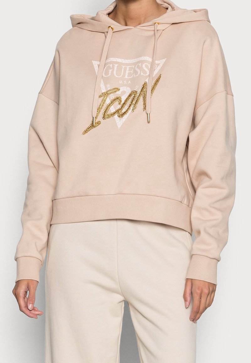 Hoodie beige con logo "Guess USA" e scritta dorata "Icon", indossato con pantaloni beige abbinati, mostrando la parte superiore del corpo e le mani.