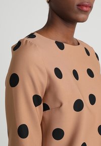 Närbild av en person som bär en beige topp med stora svarta prickar, visar axlar, nacke och läppar mot en enkel bakgrund.