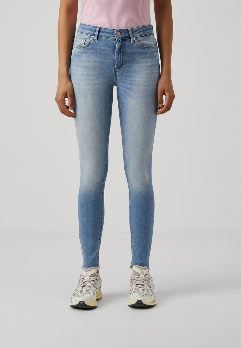 Jeans skinny de denim azul claro con efecto desvanecido, talle alto, cinco bolsillos y dobladillo deshilachado, combinados con zapatillas grises.
