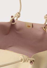 Krēmkrāsas ādas soma ar mezglotiem siksnas elementiem un maigas rozā iekšpuses oderi, aprīkota ar magnētiskās pogas aizdari un zelta krāsas metāla gredzenu.