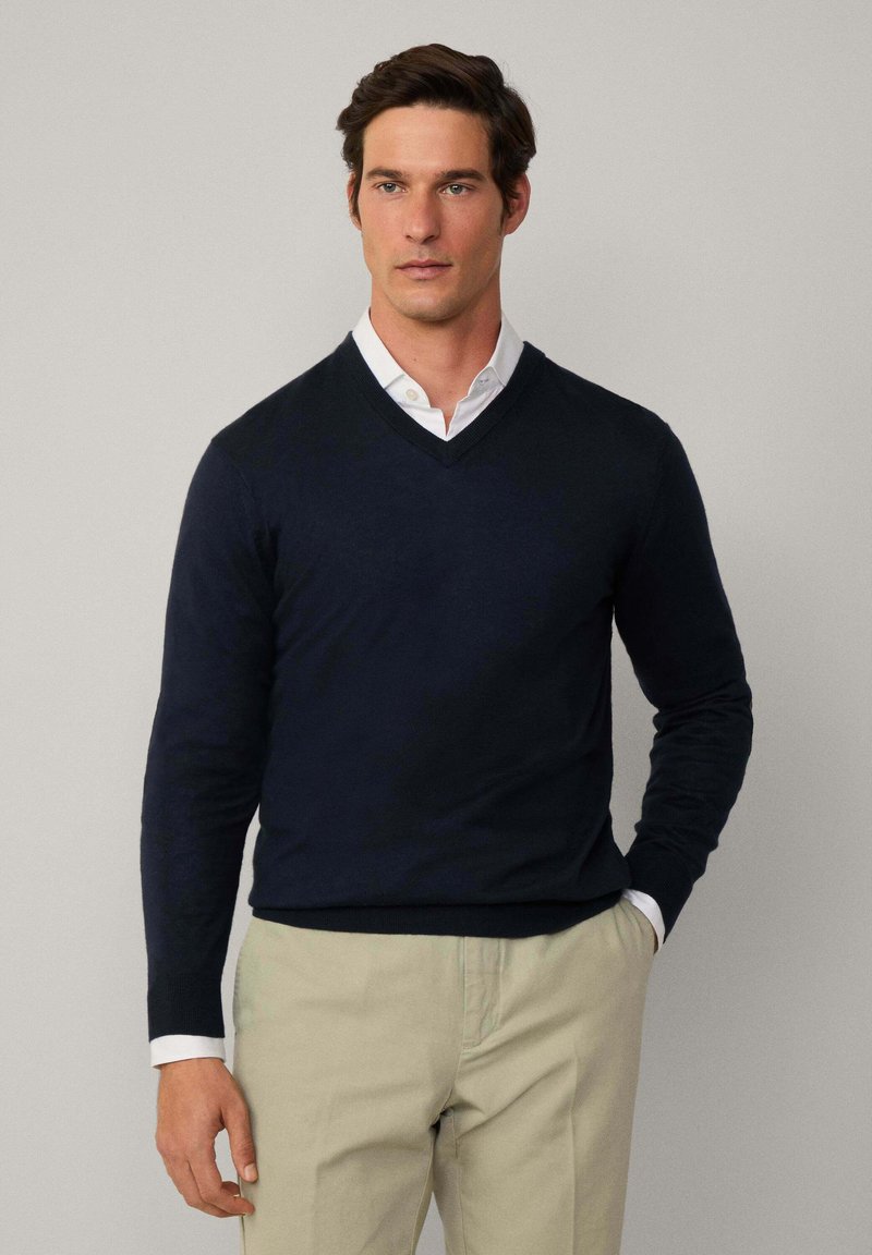 Navyblauer V-Ausschnitt-Pullover aus weichem Strickstoff, kombiniert mit einem weißen Hemd mit Kragen, beigen Hosen und einem neutralen Hintergrund.