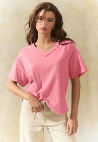 Rosa, leger geschnittenes T-Shirt mit V-Ausschnitt, kurzen Ärmeln und Brusttasche, kombiniert mit cremefarbenen, hoch taillierten Jeans. Weiche Stofftextur.