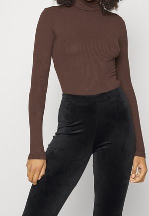 Kobieta ubrana w dopasowany brązowy prążkowany sweter z golfem i czarne, wysokowcześnione aksamitne spodnie, stojąca z ramionami luźno opuszczonymi wzdłuż ciała.