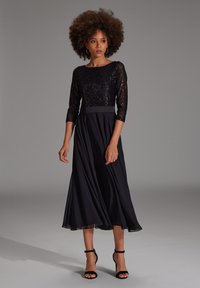 Robe noire avec un corsage pailleté, des manches 3/4, une taille cintrée et une jupe en chiffon fluide. Associée à des escarpins noirs à brides.