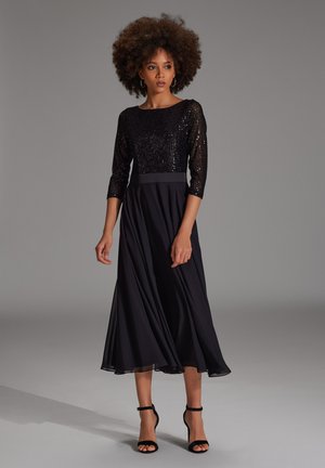 Zwarte jurk met een paillettenkorset, 3/4 mouwen, een getailleerde taille en een vloeiende chiffon rok. Gecombineerd met zwarte strapossa's.