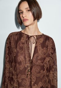 Blusa marrone con motivo paisley, design con collo a cravatta, tessuto trasparente e maniche lunghe. Presenta collane sovrapposte con pendenti decorativi.