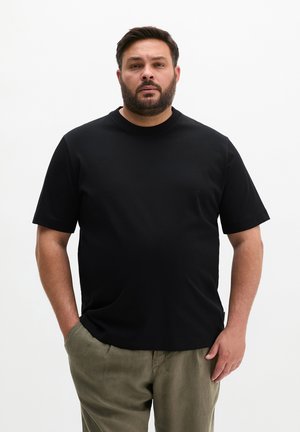 Mann mit Bart, der ein schwarzes kurzärmeliges Hemd und olivgrüne Hose trägt, steht mit einer Hand in der Tasche vor weißem Hintergrund.