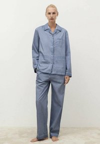 Svetlo modra pižama iz mehkega materiala, ki vključuje srajco z gumbi in ovratnikom ter žepi na prsih, v kombinaciji z ohlapnimi hlačami.