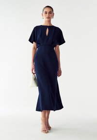 Tummansininen midi-mekko, jossa on avonainen kaula-aukko, lyhyet puffiset hihat, vyötäisillä istuva malli ja liehukehame. Asusteena pieni valkoinen käsilaukku.