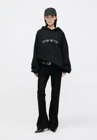 Fekete oversized kapucnis felső, kontrasztos fehér logóval, fekete harangnadrággal ésstúdiós övvel párosítva. A megjelenést egy fekete sapka és hegyes orrú cipők egészítik ki.