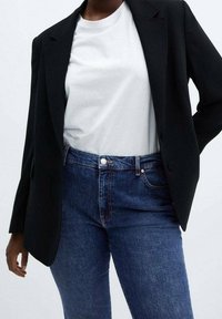 Blazer noir sur mesure porté sur un t-shirt blanc, associé à un jean bleu foncé. Les détails notables incluent des surpiqûres visibles sur le jean et les revers du blazer.