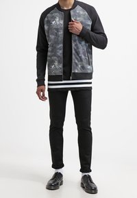 Chaqueta bomber negra con un frente texturizado en gris y azul; puños acanalados; superpuesta sobre una camiseta negra; combinada con jeans y zapatos negros.