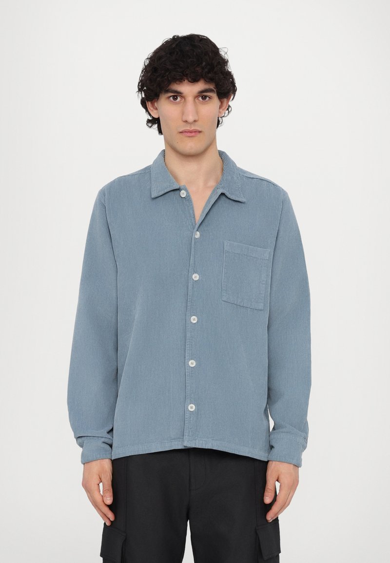 Light blue corduroy shirt with a collar, button closure, long sleeves, and a chest pocket. Textured fabric with vertical stripes.