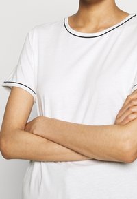 Camiseta blanca de manga corta con cuello redondo y ribete negro en el cuello y las mangas. La tela parece suave con una textura lisa.