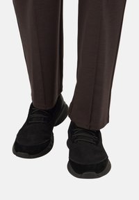 Osoba v tmavohnedých nohaviciach a čiernych šnurovacích teniskách na jednoduchej bielej ploche.