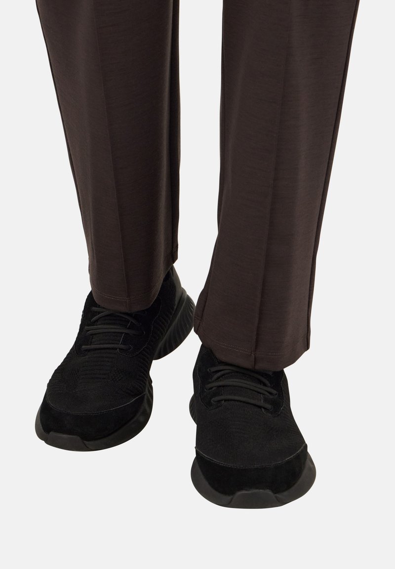 Osoba v tmavohnedých nohaviciach a čiernych šnurovacích teniskách na jednoduchej bielej ploche.