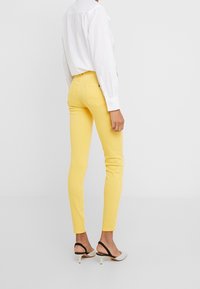 Jeans ajustados amarillos de mezclilla, con una cintura alta y dos bolsillos traseros. Combinados con una camisa blanca de manga larga y sandalias negras de tacón.