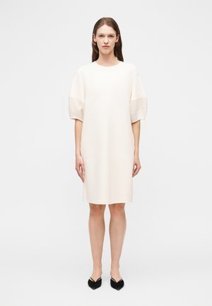 Žena stojaca čelom vpred v voľných krémových šatách dĺžky po kolená s polovičnými rukávmi a čiernymi topánkami so špicatou špičkou na bielom pozadí.