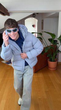 Lys grå dunjakke med hette, kombinert med en marineblå hettegenser og lyse denimjeans. Blå solbriller; innendørs setting med planter.