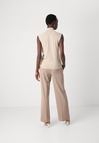 Svetlo bež brezrokavnik s visokim ovratnikom, v kombinaciji s svetlo rjavimi hlačami s črtami. Gladka tekstura, prilega se telesu in subtilni detajli.