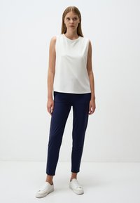 Witte mouwloze top in een gladde stof, gecombineerd met marineblauwe op maat gemaakte broek. Model draagt witte sneakers. Effen achtergrond.