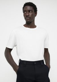 T-shirt branco de mangas curtas feito de algodão macio, combinado com calças escuras ajustadas que possuem fecho de botão e bolsos. Textura suave.