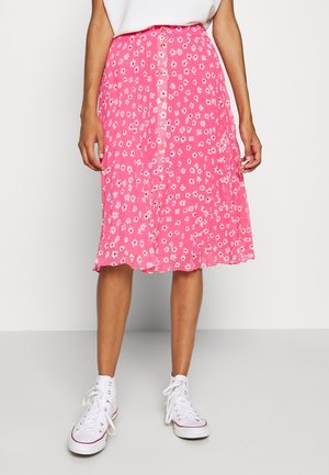 Falda rosa con estampado floral hasta la rodilla, con botones en el centro, combinada con zapatillas blancas altas y camiseta blanca, de pie sobre suelo gris.