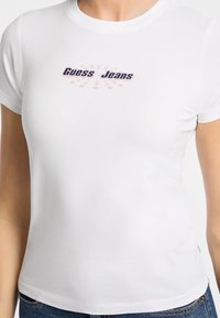 Valkoinen istuva t-paita, joka on valmistettu sileästä kankaasta. Tässä paidassa on sininen "Guess Jeans" -logo, jossa on vaaleanpunaisia pyöreitä korostuksia. Lyhyet hihat ja pyöreä kaula-aukko.