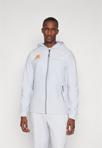 Šviesiai pilkas zip-up džemperis su kapišonu. Turi oranžinius logotipo akcentus ir elastingas rankoves, pagamintas iš švelnios, lengvos medžiagos.