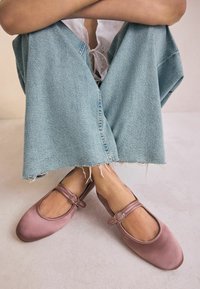 Scarpe Mary Jane in raso rosa con punta rotonda, dotate di due fettucce con fibbie circolari, abbinate a pantaloni in denim blu chiaro con orli sfilacciati.