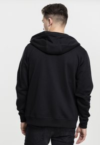 Hoodie noir en matériau texturé, avec une grande capuche, des poignets et un ourlet côtelés. Design simple, sans logos ni motifs visibles.