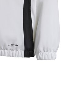 Witte jas met zwarte zijpanelen, voorzien van een textuurafwerking en elastische manchetten. "AEROREADY" logo aan de onderzijde links.